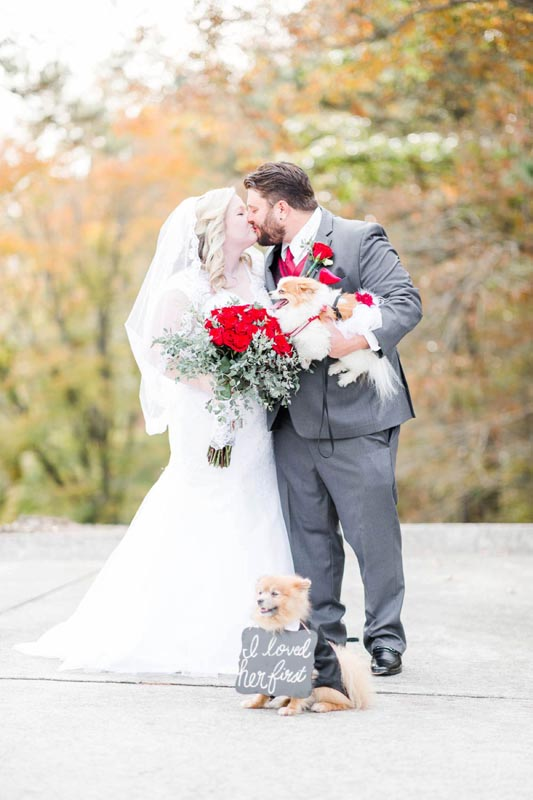 smiling bride and groom kissing while holding flowers and a puppy. A small puppy sits at their feet with the sign "I loved her first".